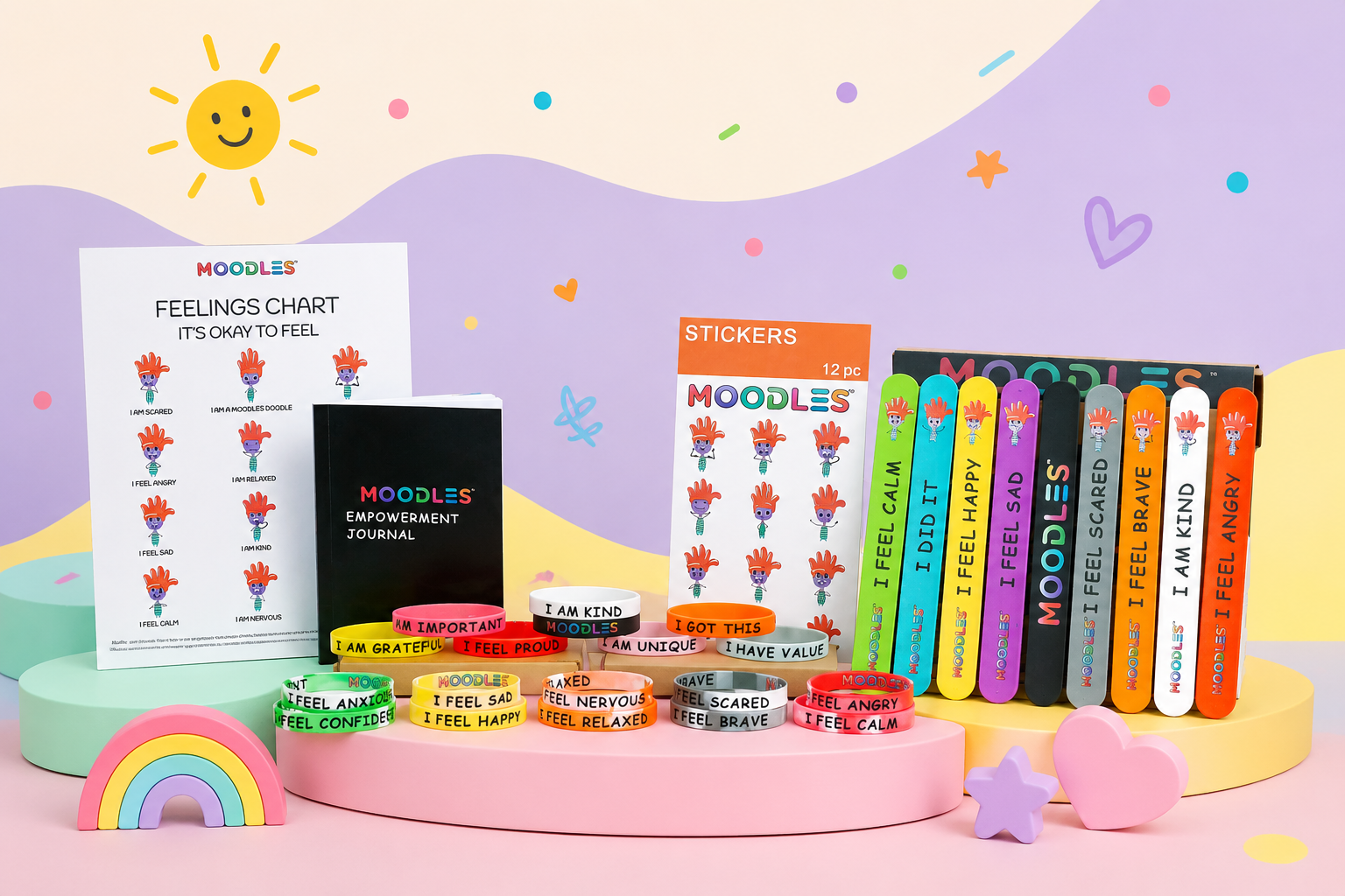A colorful Moodles stationery and self-care gift set arranged on pastel display pedestals, including a feelings chart, affirmation journal, sticker sheet, wristbands, and colorful pens or markers, with a sun, rainbow, and heart illustrations in the background.
