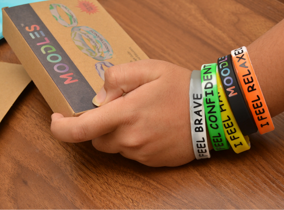 Hand wearing colorful wristbands with text, holding a box on a wooden surface