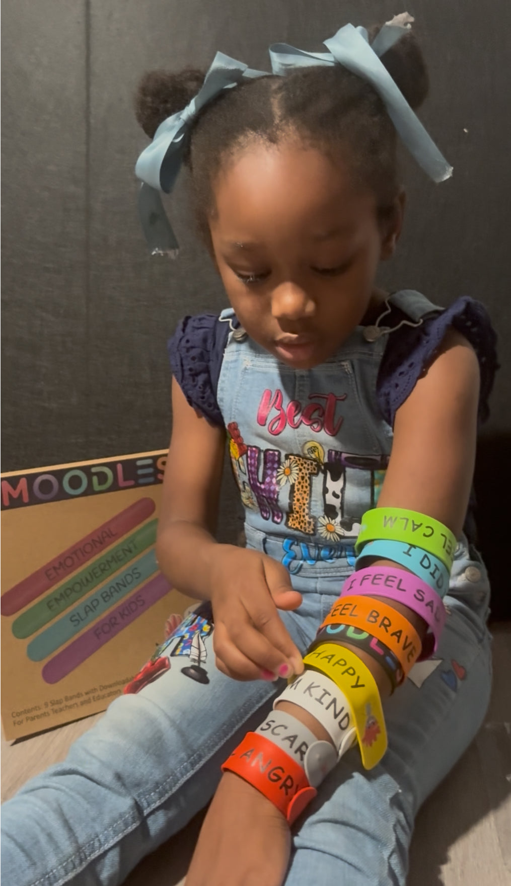 Child wearing colorful bracelets with messages, sitting on the floor with a box in the background.